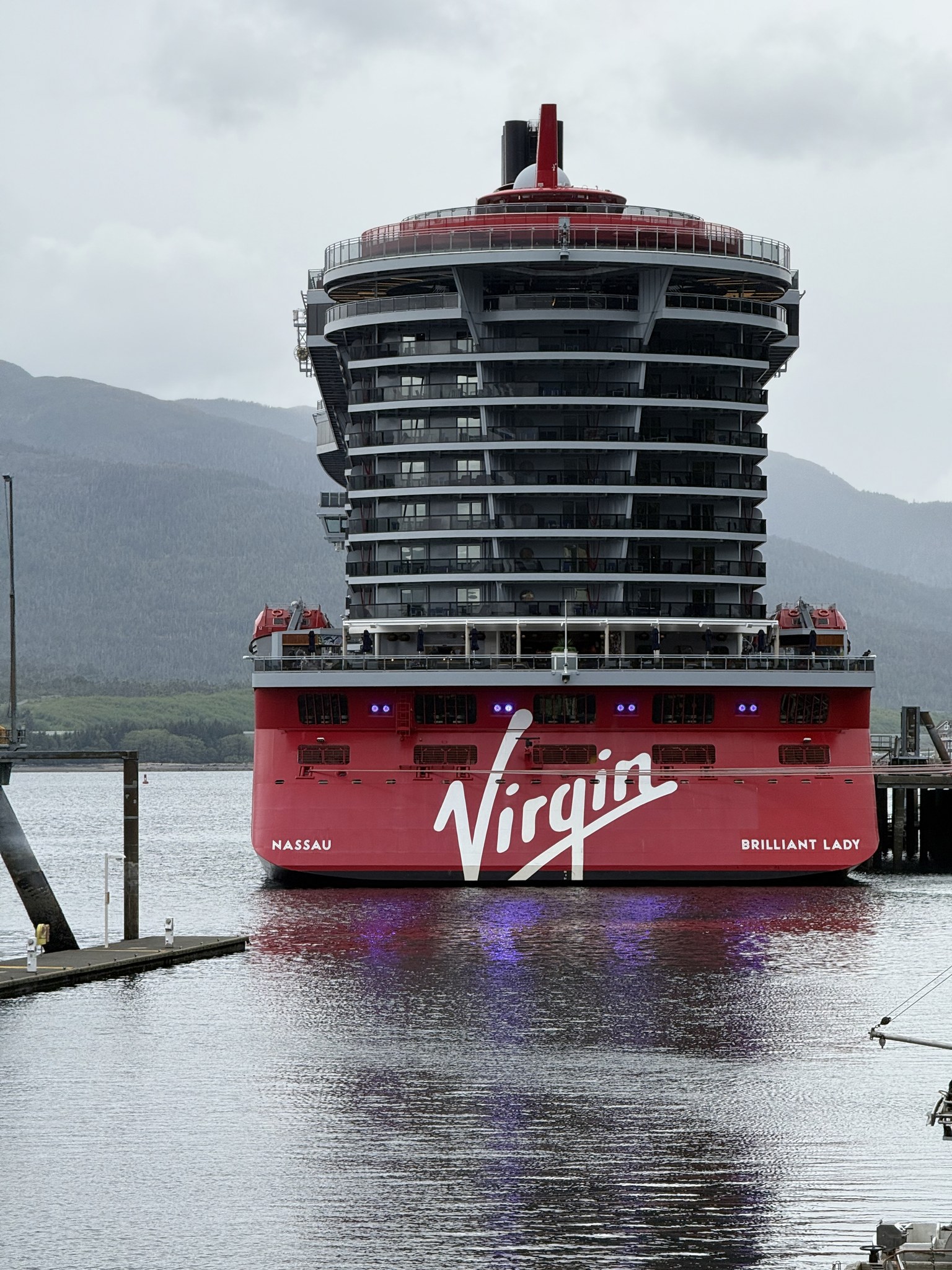Brilliant Lady’s red Virgin stern alongside in Alaska with mountains in the background